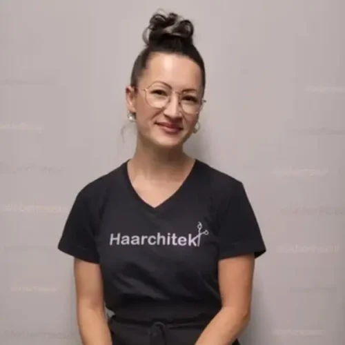 Frau mit Brille in einem Haarchitek-Shirt vor grauem Hintergrund.