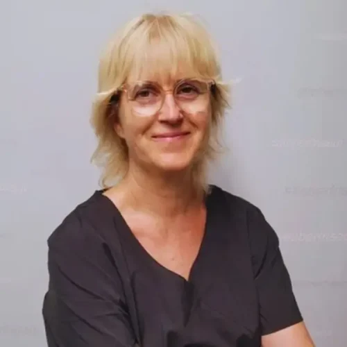 Eine lächelnde Person mit kurzen blonden Haaren und Brille, die ein dunkles Oberteil trägt.